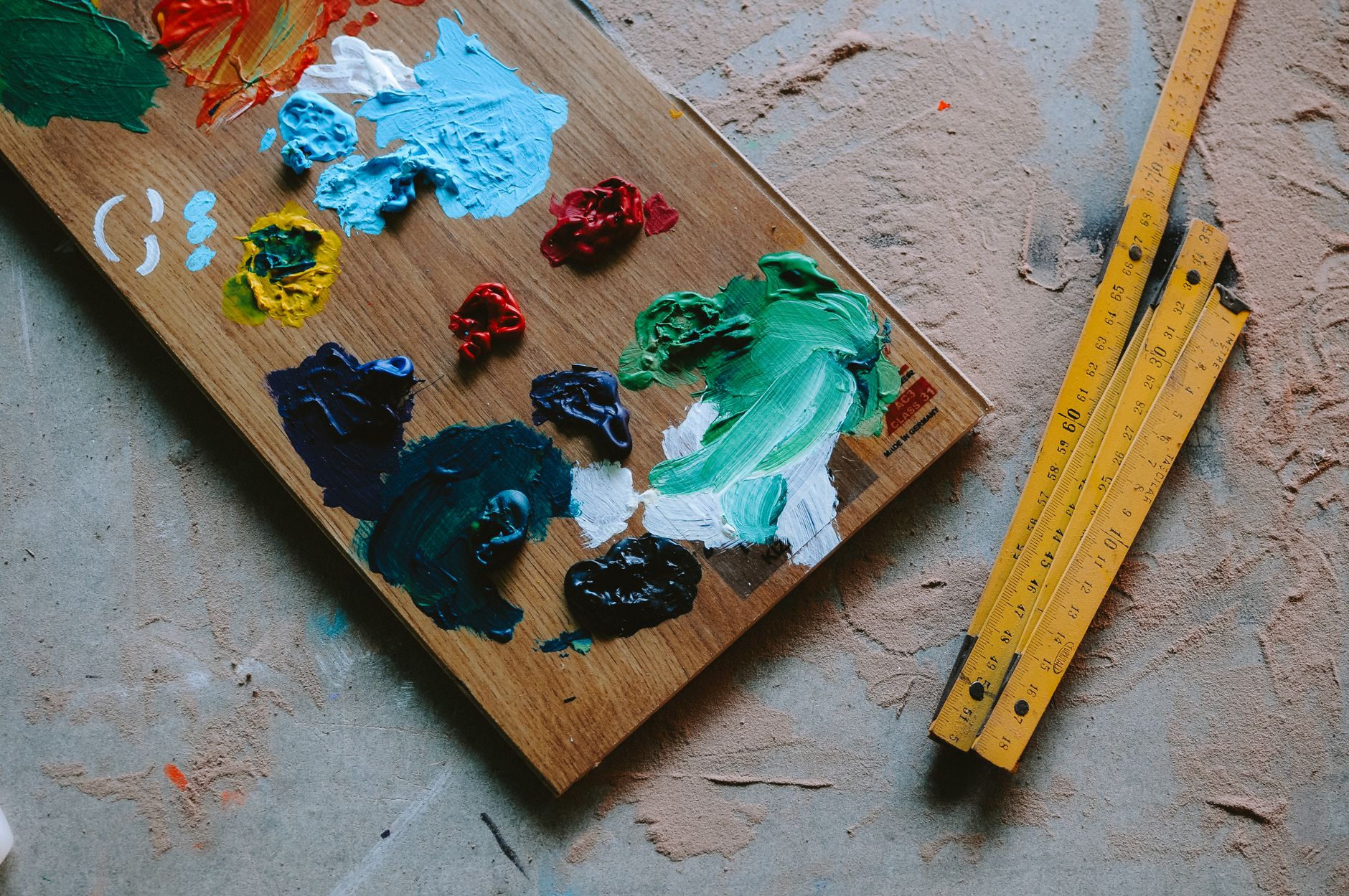 Colorful oil paints on a wooden palette beside wooden measuring tapes on a textured floor in Carlsbad, CA